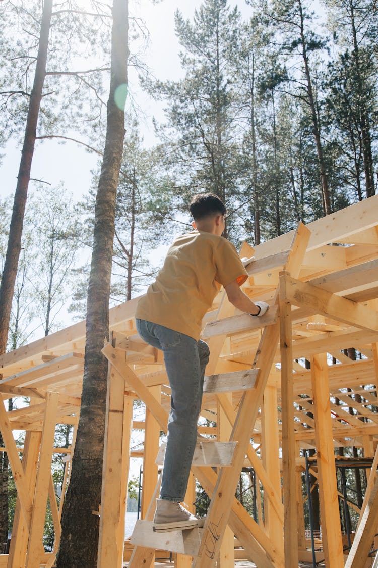 A Man In A Yellow Shirt And Denim Pants Climbing A Wooden Ladder