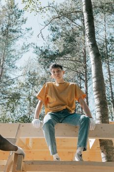 A young adult male in a yellow shirt and jeans sitting on a wooden structure amid tall trees.
