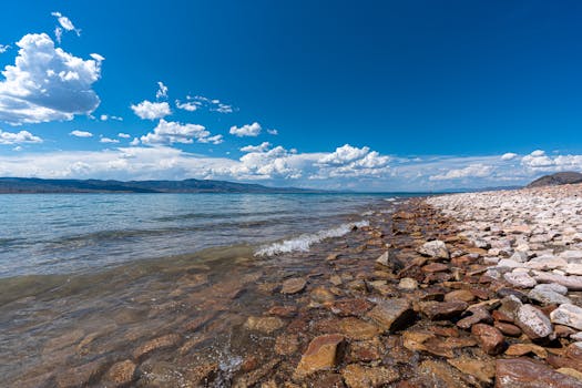 Crystal clear waves washing over rocky shore under a vibrant blue sky and fluffy clouds.