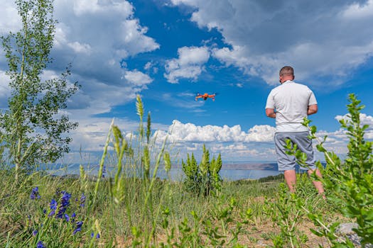 A man in a white shirt flies a drone over a scenic outdoor landscape with blue skies and green grass.