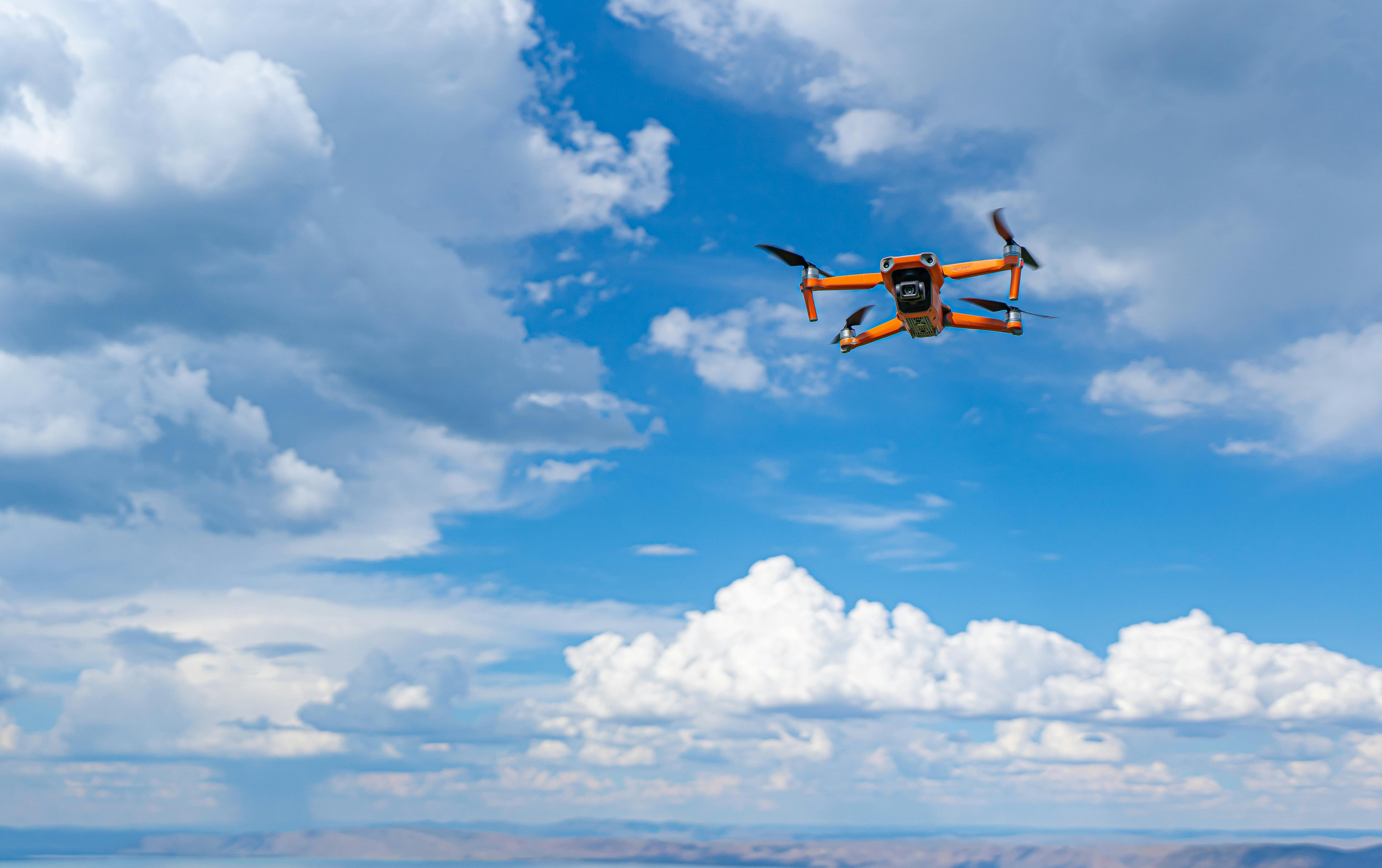 orange and black drone flying under blue sky and white clouds