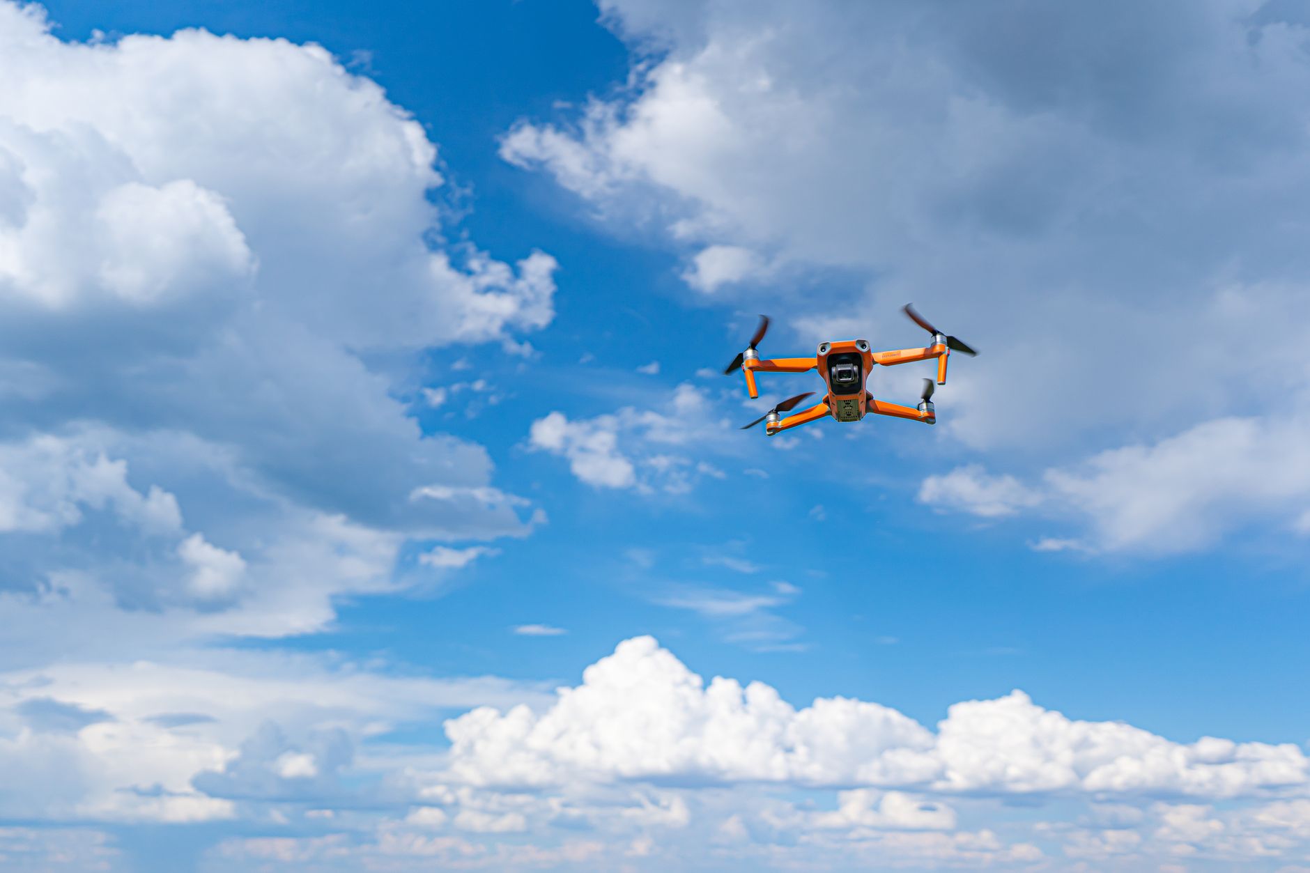 Aerial view of an orange drone flying against a bright blue sky with fluffy clouds.