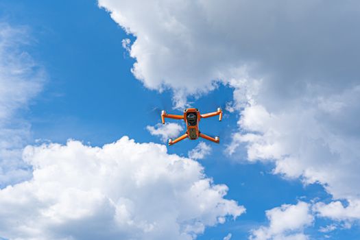 An orange drone captured mid-flight against a clear blue sky with fluffy clouds, embodying modern technology.