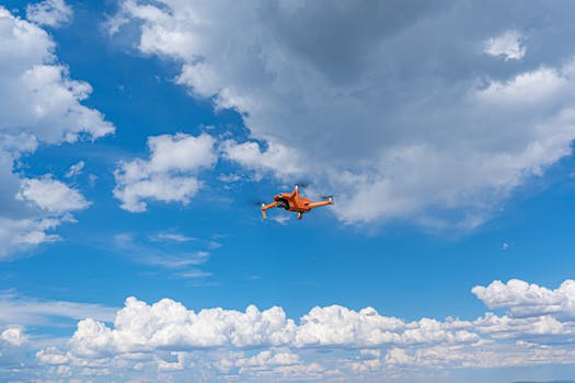 An orange drone flying high amidst a vibrant blue sky with scattered clouds.