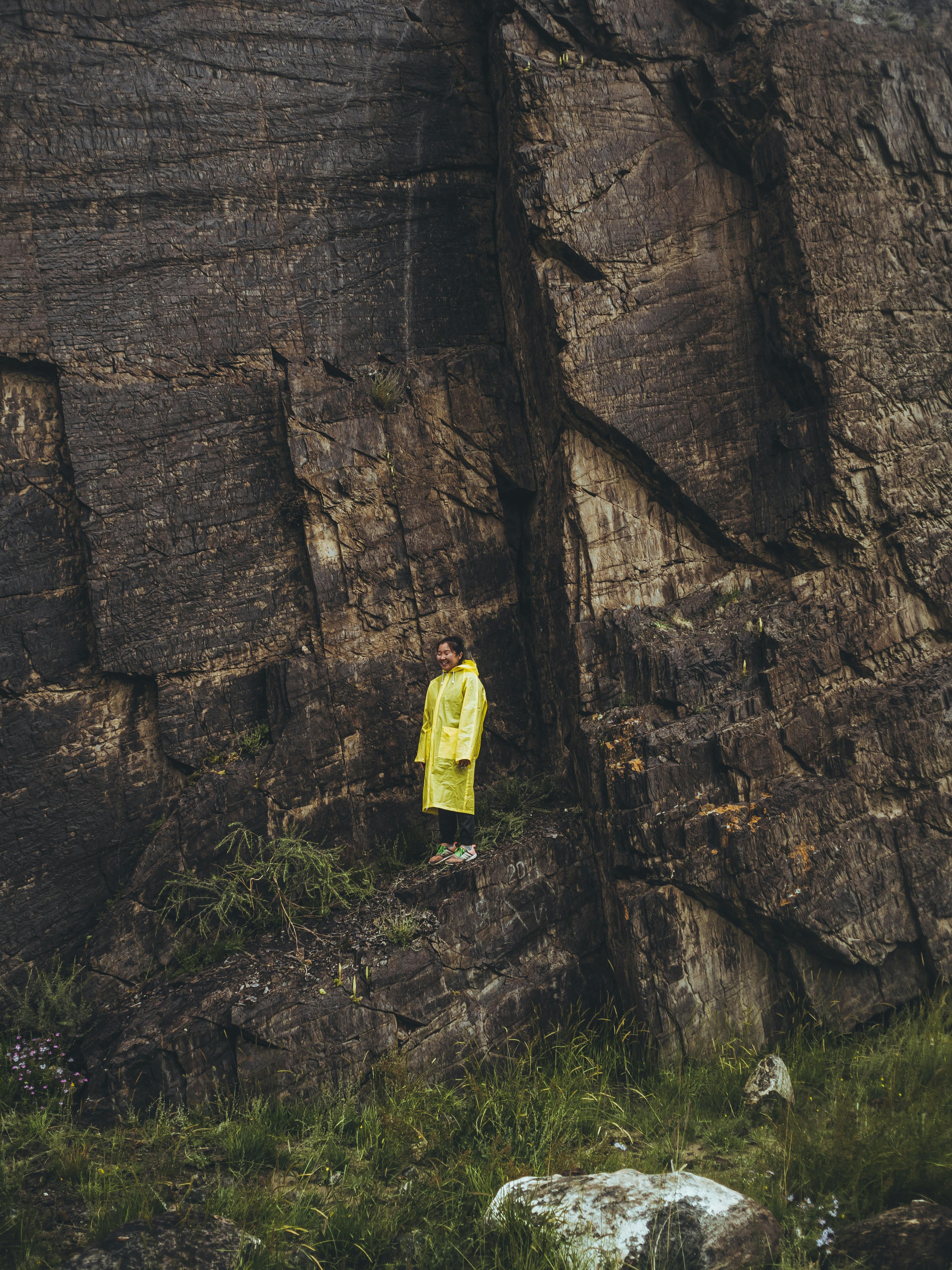 A person in a yellow raincoat stands by a rugged rock wall in the Altai Republic, showcasing nature's raw beauty.
