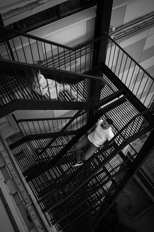 Grayscale Photo of a Man and Woman Sitting on Metal Staircase of a Building