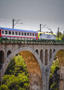 Colorful train crossing a historic stone viaduct bridge surrounded by lush green landscape.