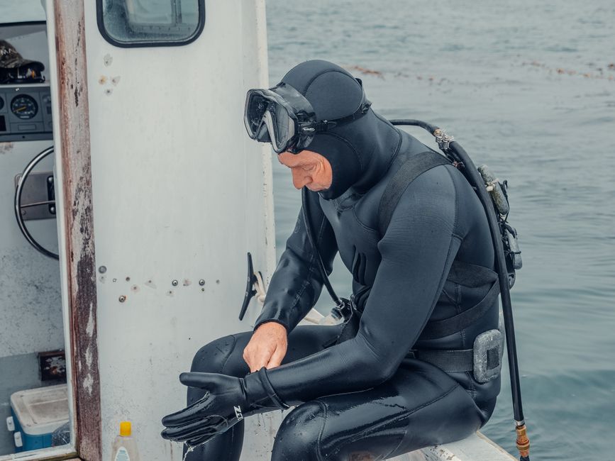 A male diver sitting on a boat, wearing a wetsuit and gloves, preparing his gear for a dive.