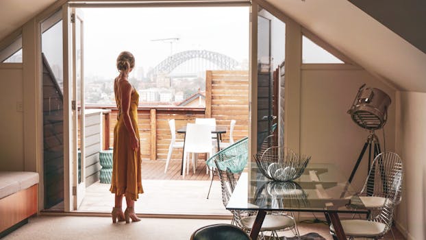 Woman in elegant dress looks out at Sydney Harbour Bridge from modern apartment balcony.