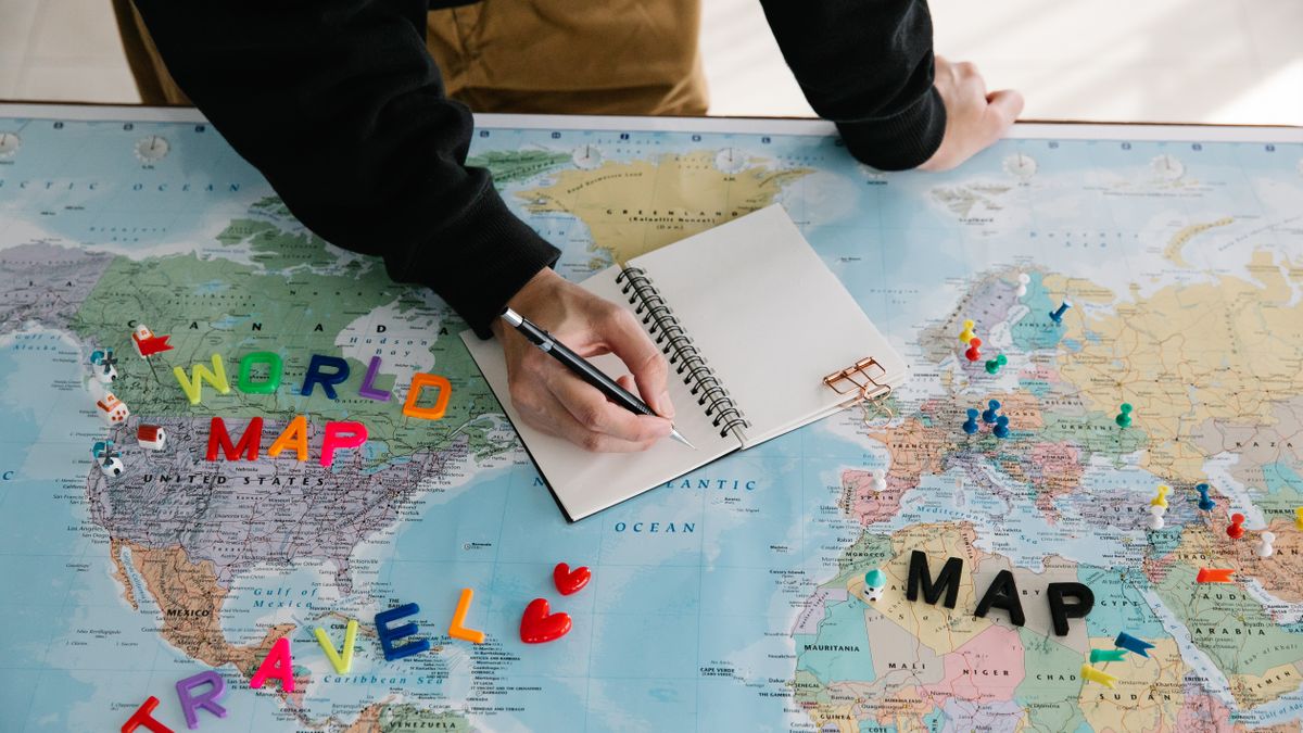 A person writing in a notebook on a world map, planning a travel journey with colorful markers and a pen.