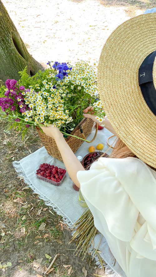 Free A woman decorates a picnic basket with flowers beneath a tree, surrounded by fruits. Stock Photo