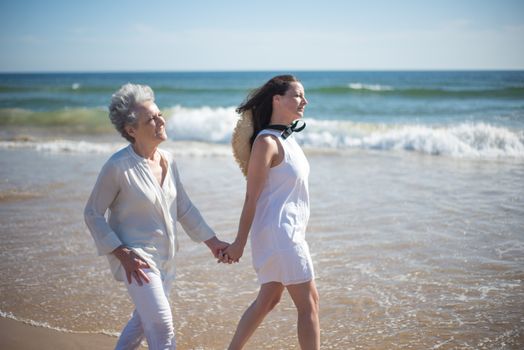 A senior woman and her daughter holding hands while walking along a sunny beach in Portugal.