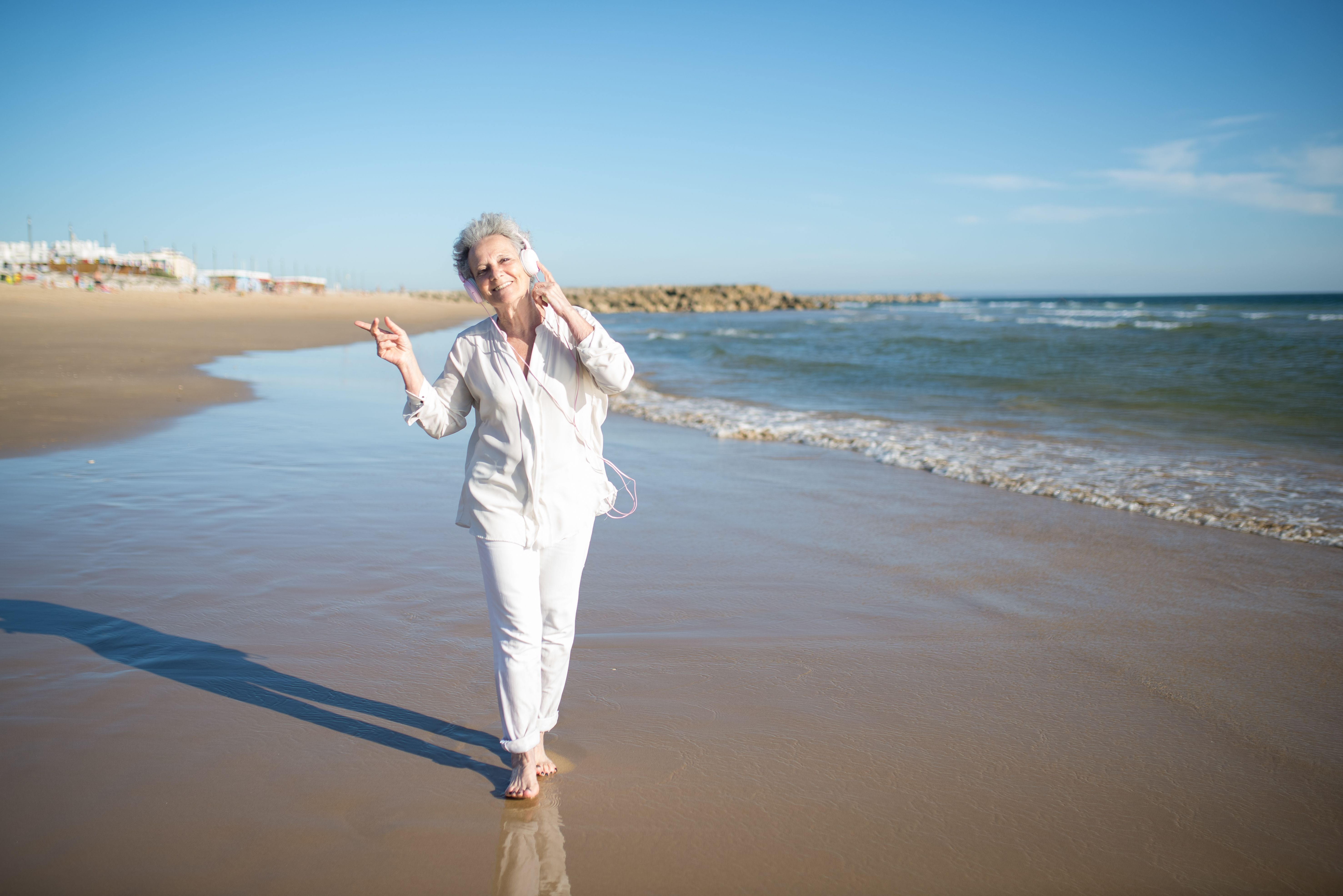 Elderly woman in white enjoying music with headphones on a sunny Portuguese beach.