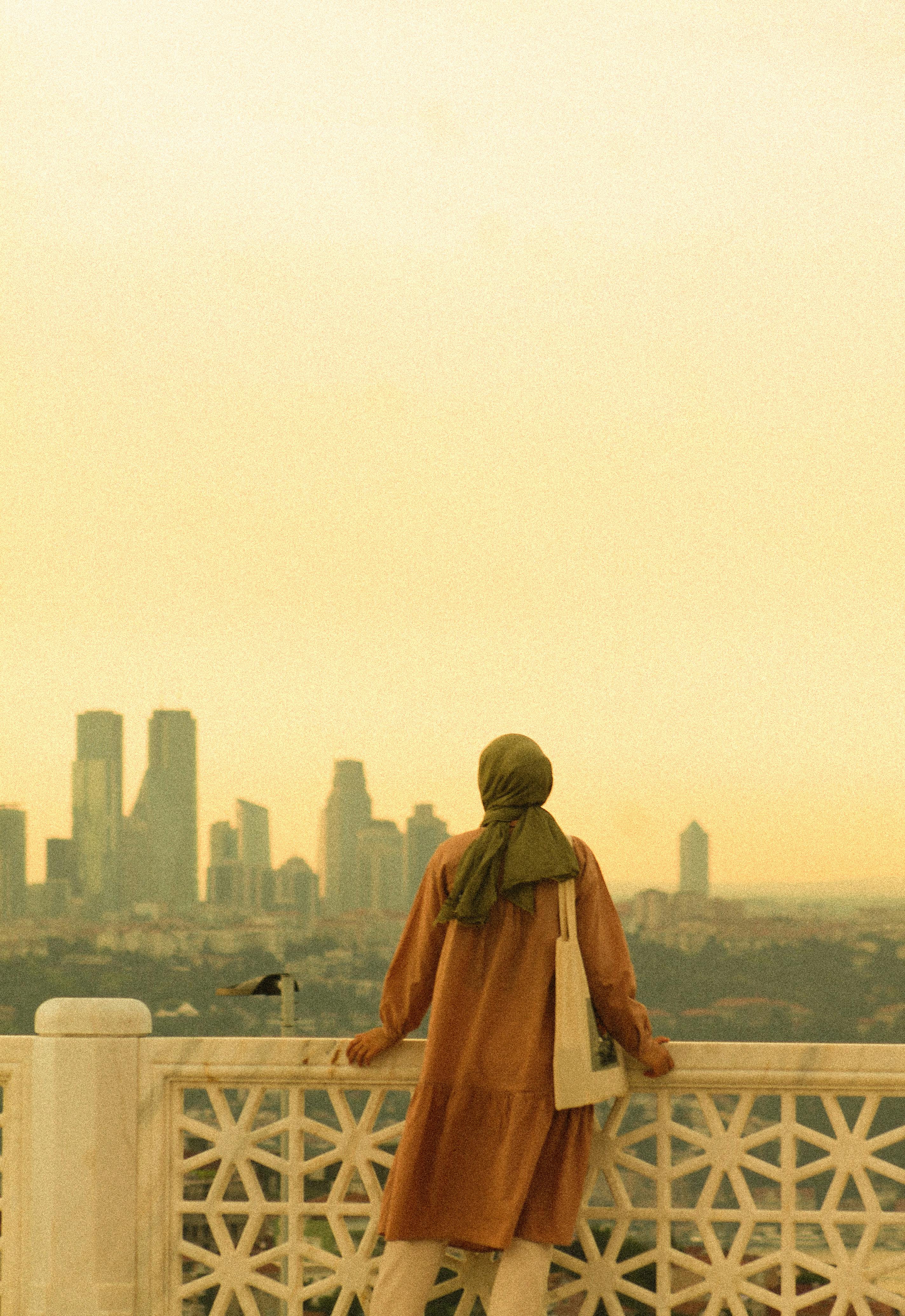 A woman in a brown dress and hijab gazes at the city skyline during sunset, creating a serene moment.