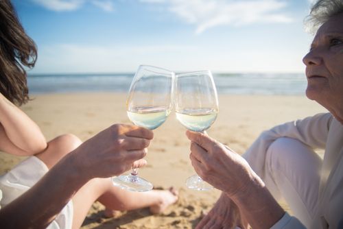 Free Two women clink wine glasses on a sandy Portuguese beach, enjoying a sunny day. Stock Photo