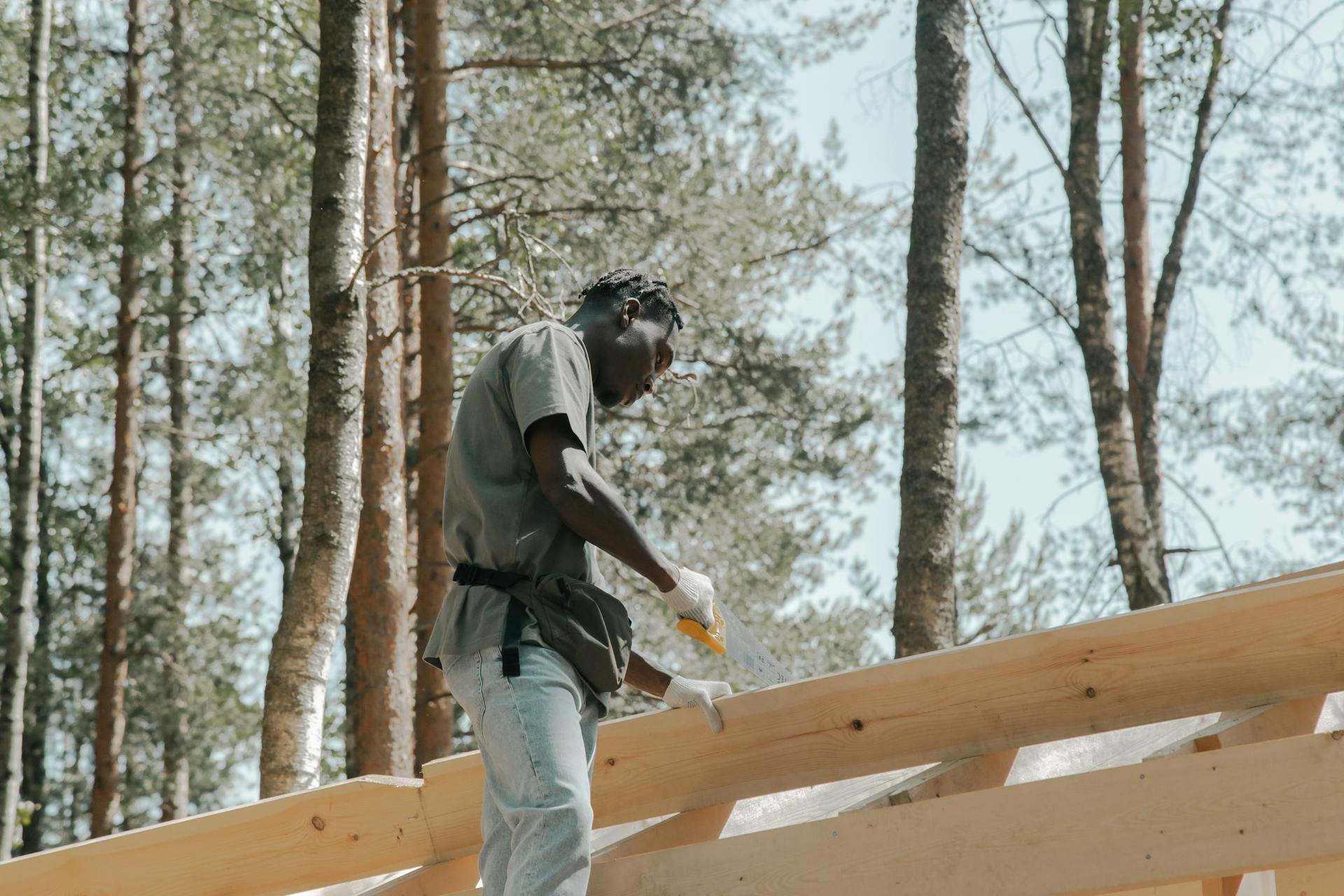 A construction worker sawing wood on a building project outdoors in a forest setting.