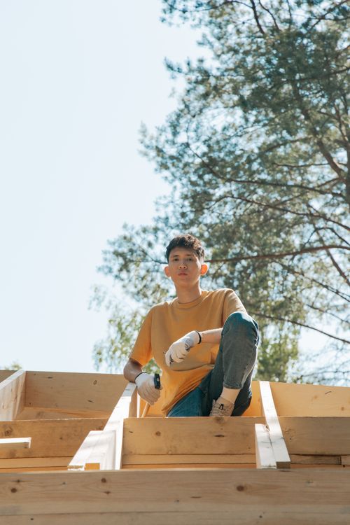 Free A young man wearing gloves works on a wooden structure outdoors, emphasizing craftsmanship. Stock Photo