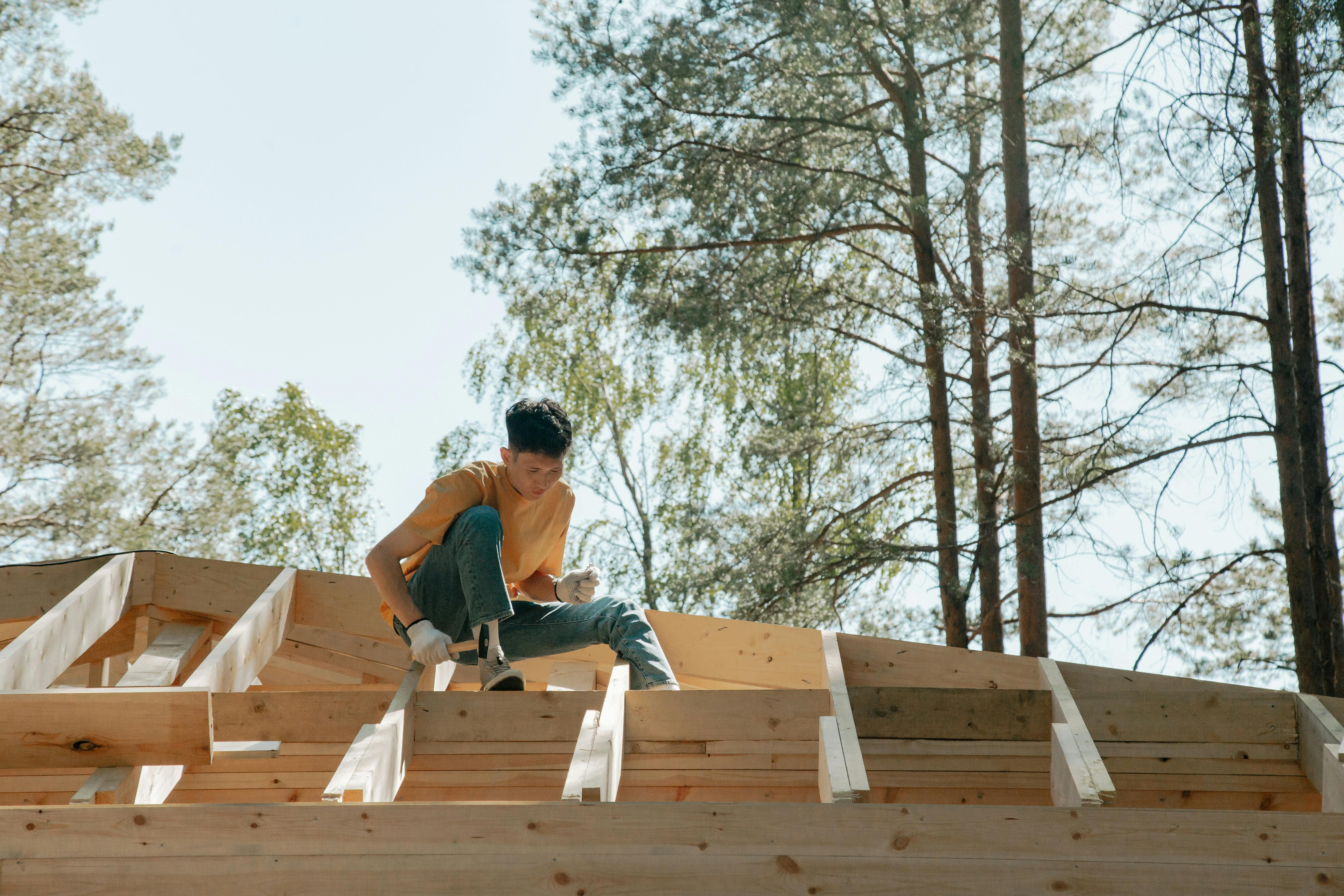 A construction worker skillfully building a wooden roof framework surrounded by tall trees.