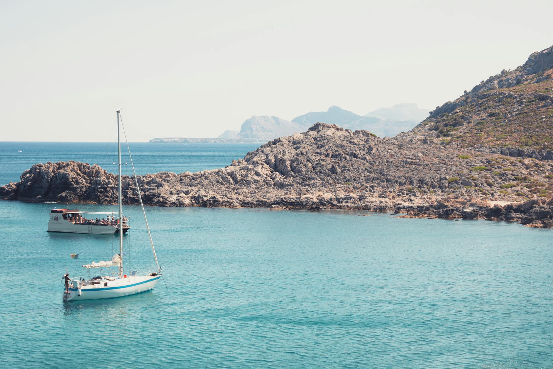 Explore the serene beauty of Rhodes with boats sailing by rocky shores under a clear sky.