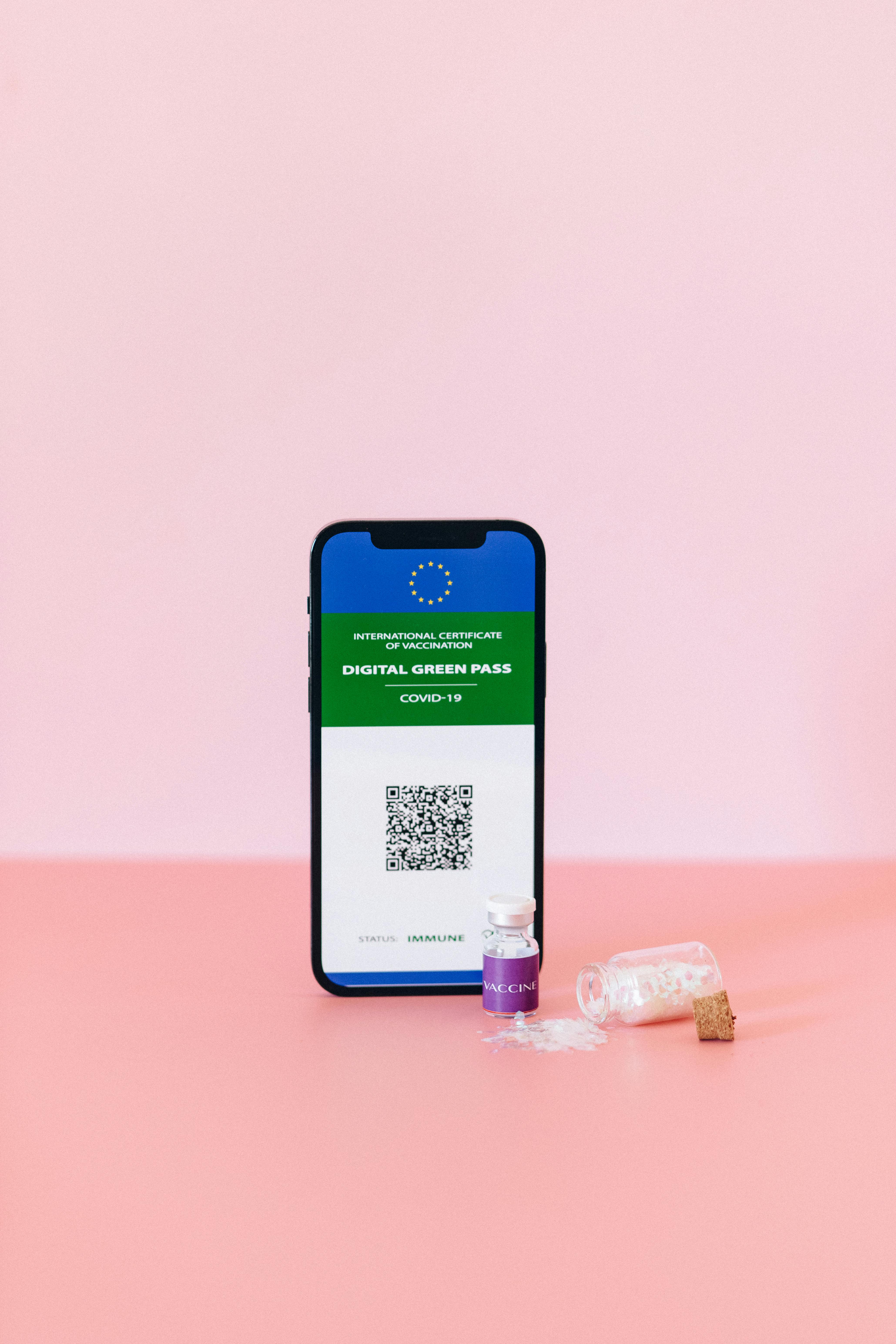 Smartphone displaying digital green pass with a vaccine vial on a pastel pink surface, studio shot.