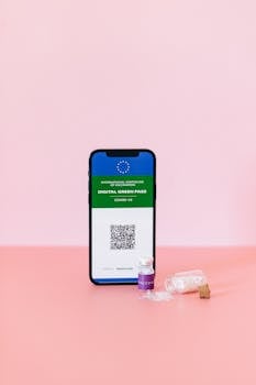 Smartphone displaying digital green pass with a vaccine vial on a pastel pink surface, studio shot.