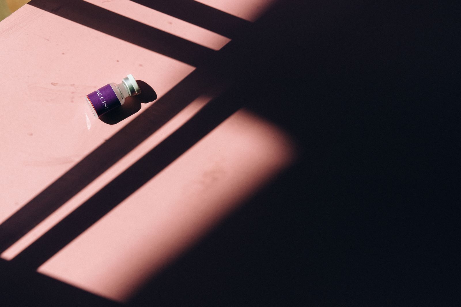 A small glass vial with a purple label lying on a pink surface in sharp window shadows