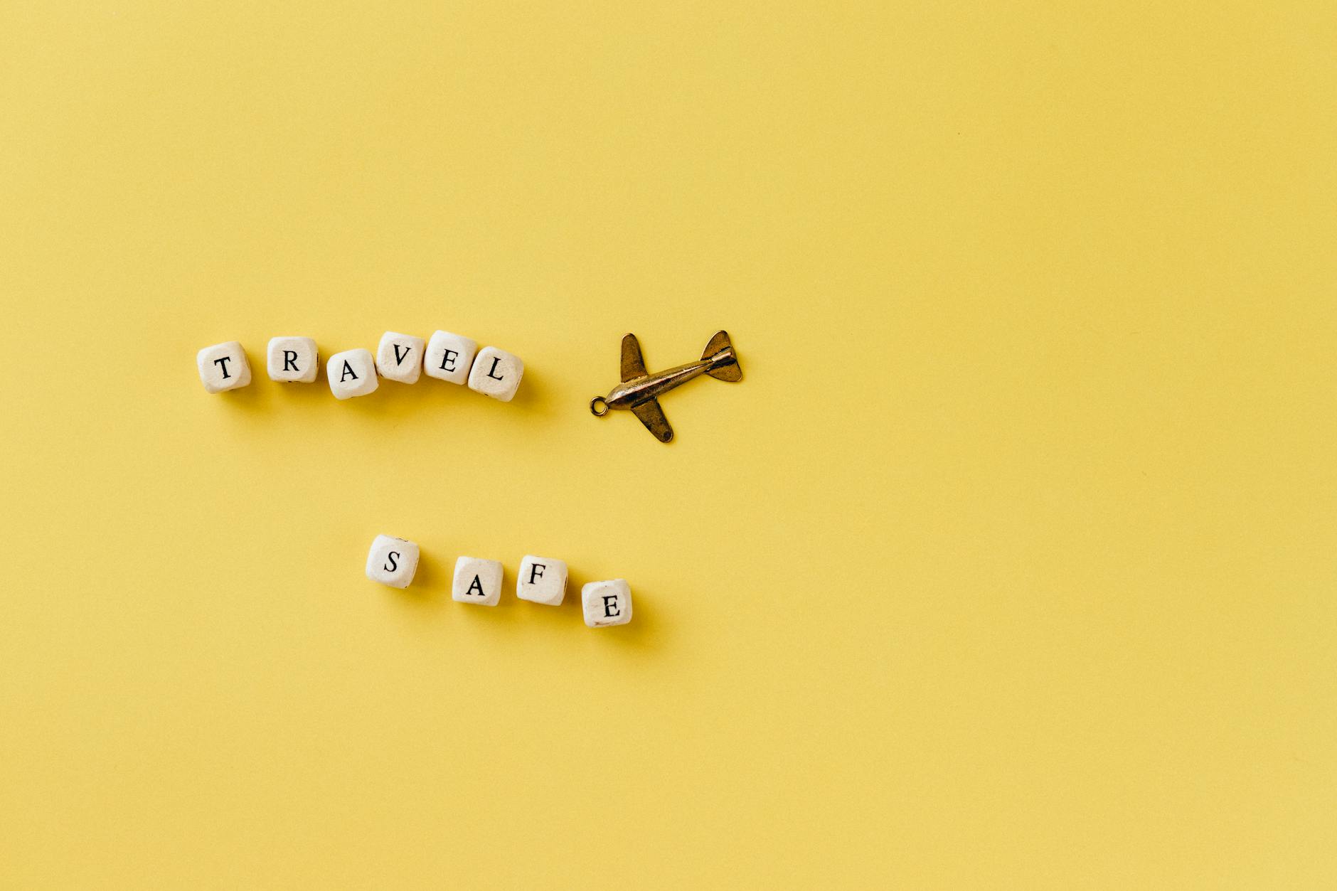Flat lay of 'Travel Safe' text with airplane pendant on yellow background, evoking travel and safety concepts.