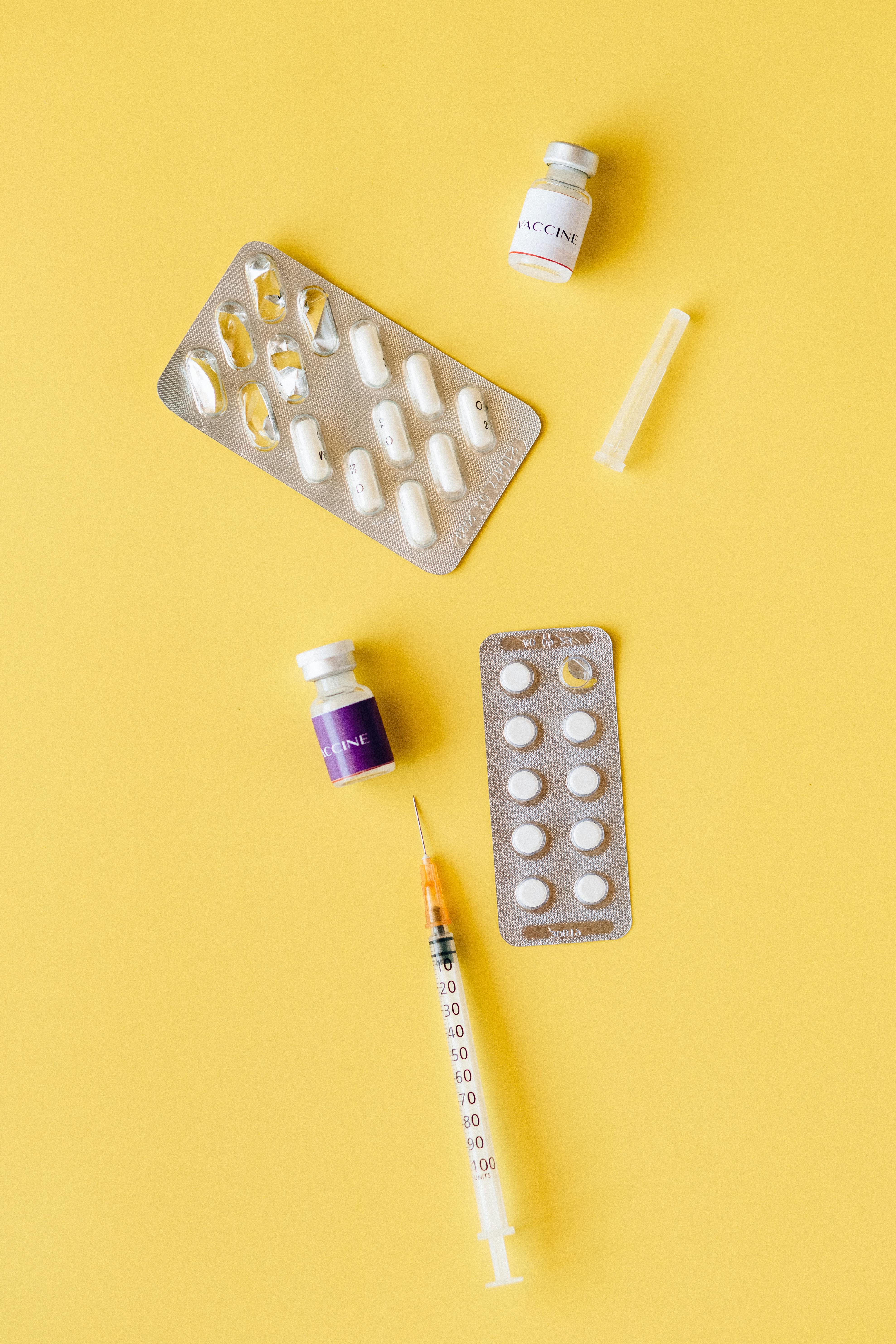 Flat lay of blister packs, vials, and syringe on yellow surface representing medical treatment.