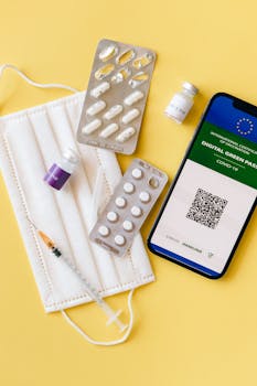 Flat lay of pandemic essentials: mask, vaccine vials, digital pass, medical blister packs, and syringe on a yellow background.