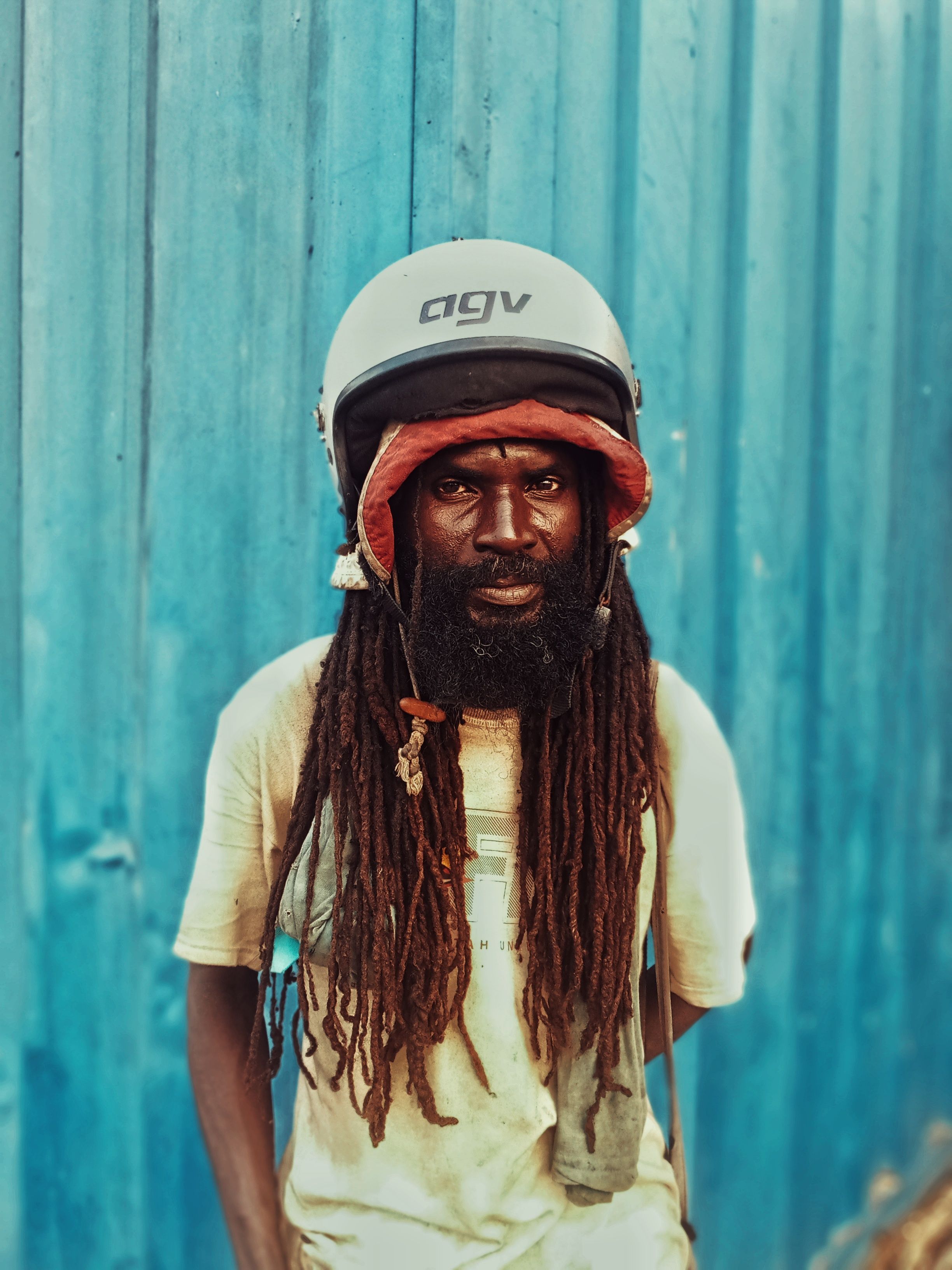 A man with dreadlocks wearing a helmet stands before a blue wall, exuding confidence.