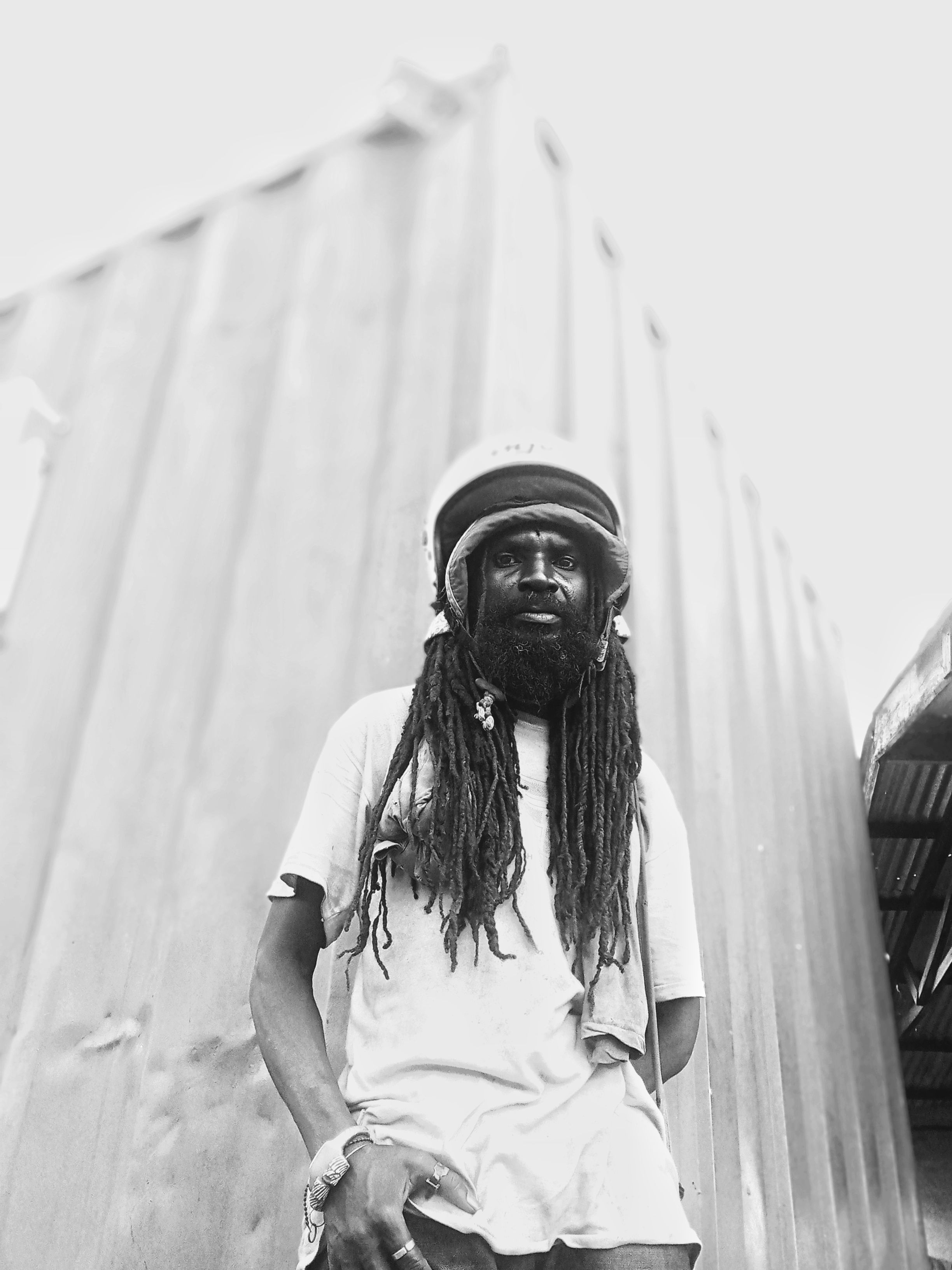 Low angle black and white photo of a man with dreadlocks wearing a helmet, standing outdoors.