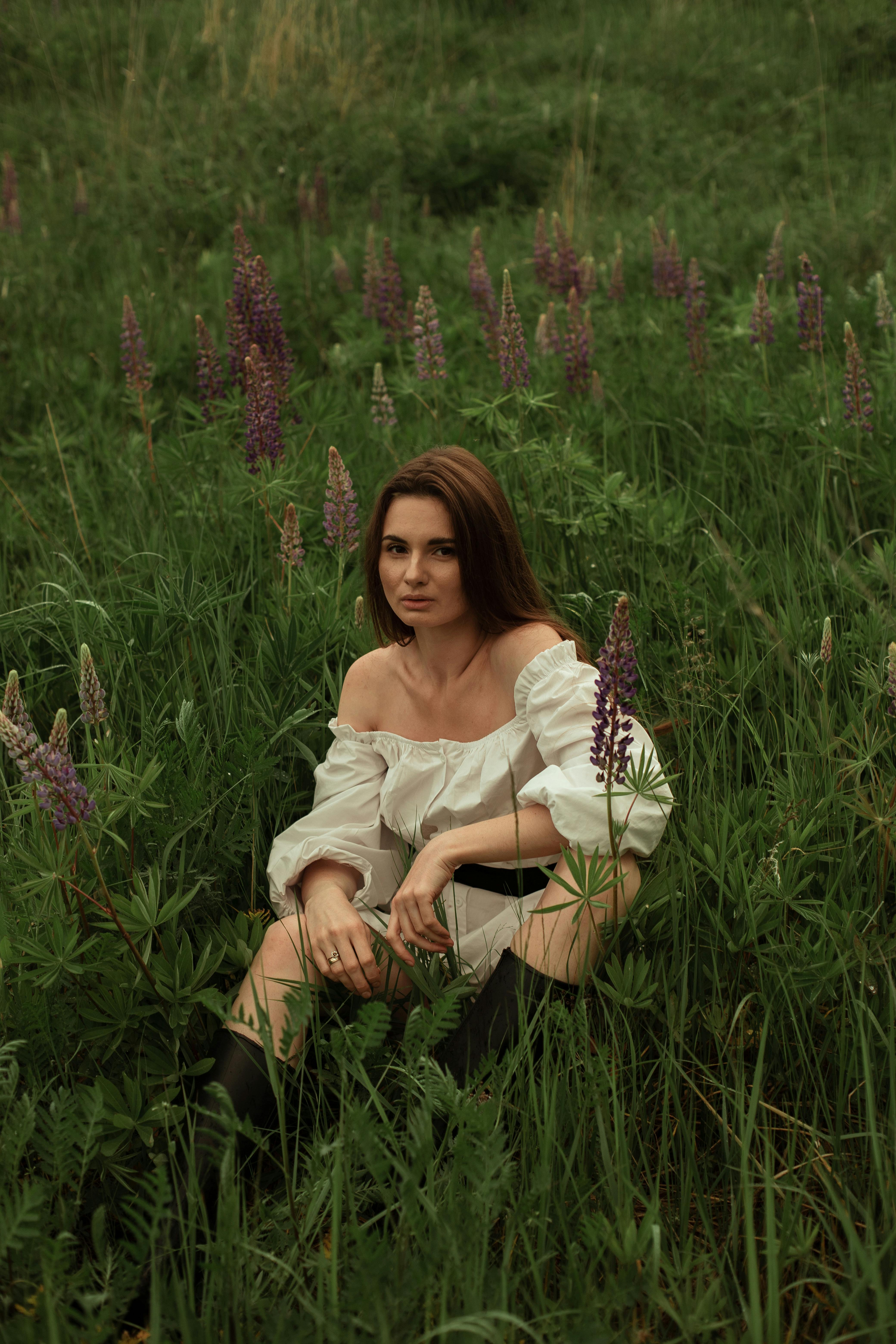 Woman sitting in lush green field surrounded by lupines, exuding calmness with an off-shoulder white dress.