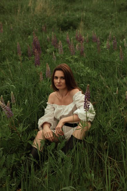 Woman sitting in lush green field surrounded by lupines, exuding calmness with an off-shoulder white dress.
