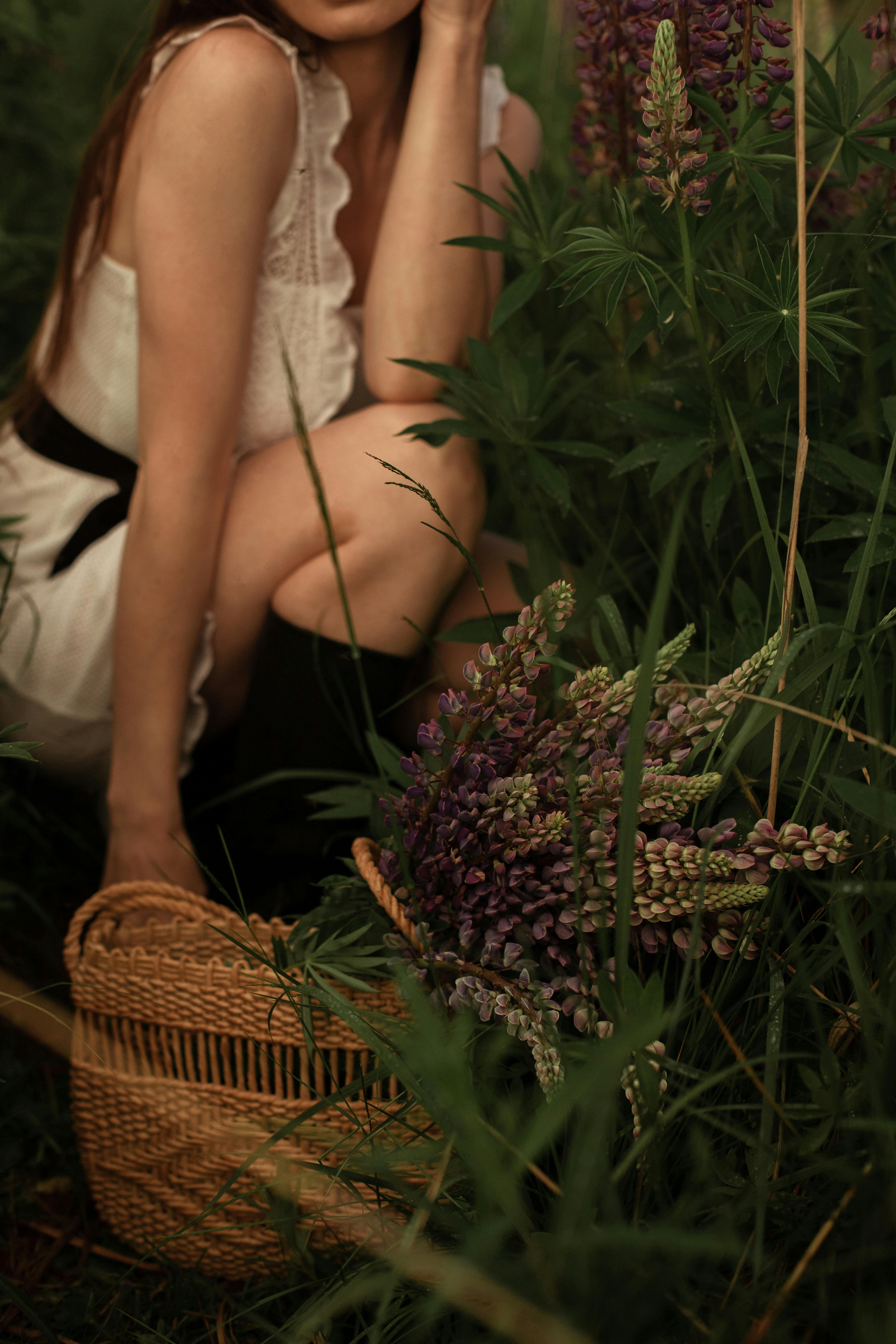 A stylish woman crouches with a basket of lupines in a lush, outdoor setting.