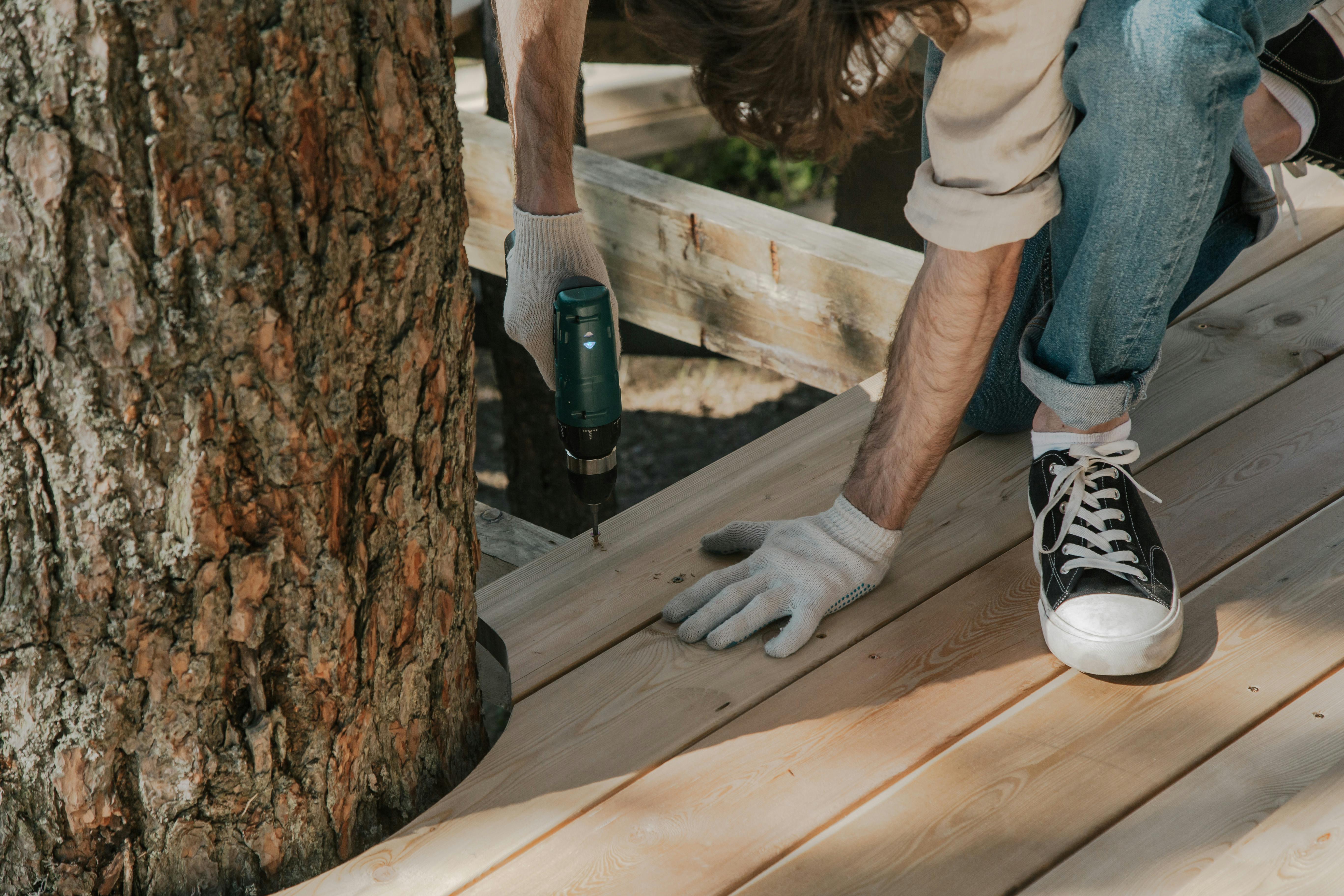 Worker drilling wooden planks on deck beside tree using power tool. DIY construction project.