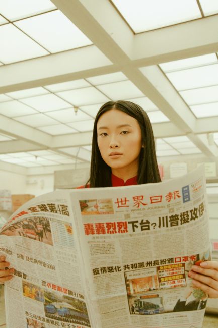 Portrait of an adult woman holding a Chinese newspaper indoors, looking serious.