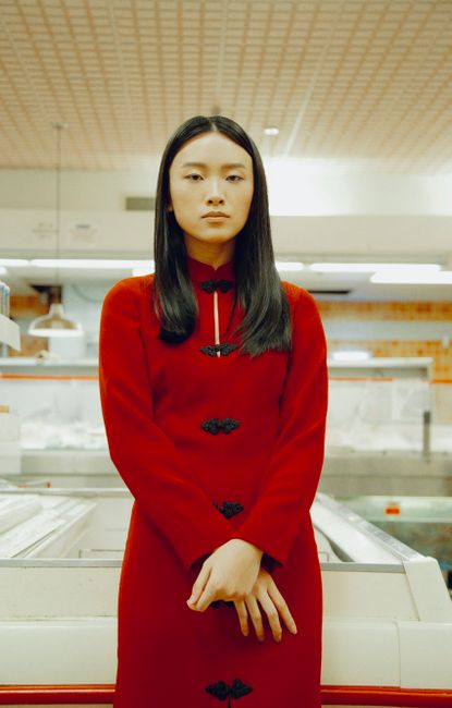 Portrait of a poised Asian woman in a striking red dress standing confidently indoors.
