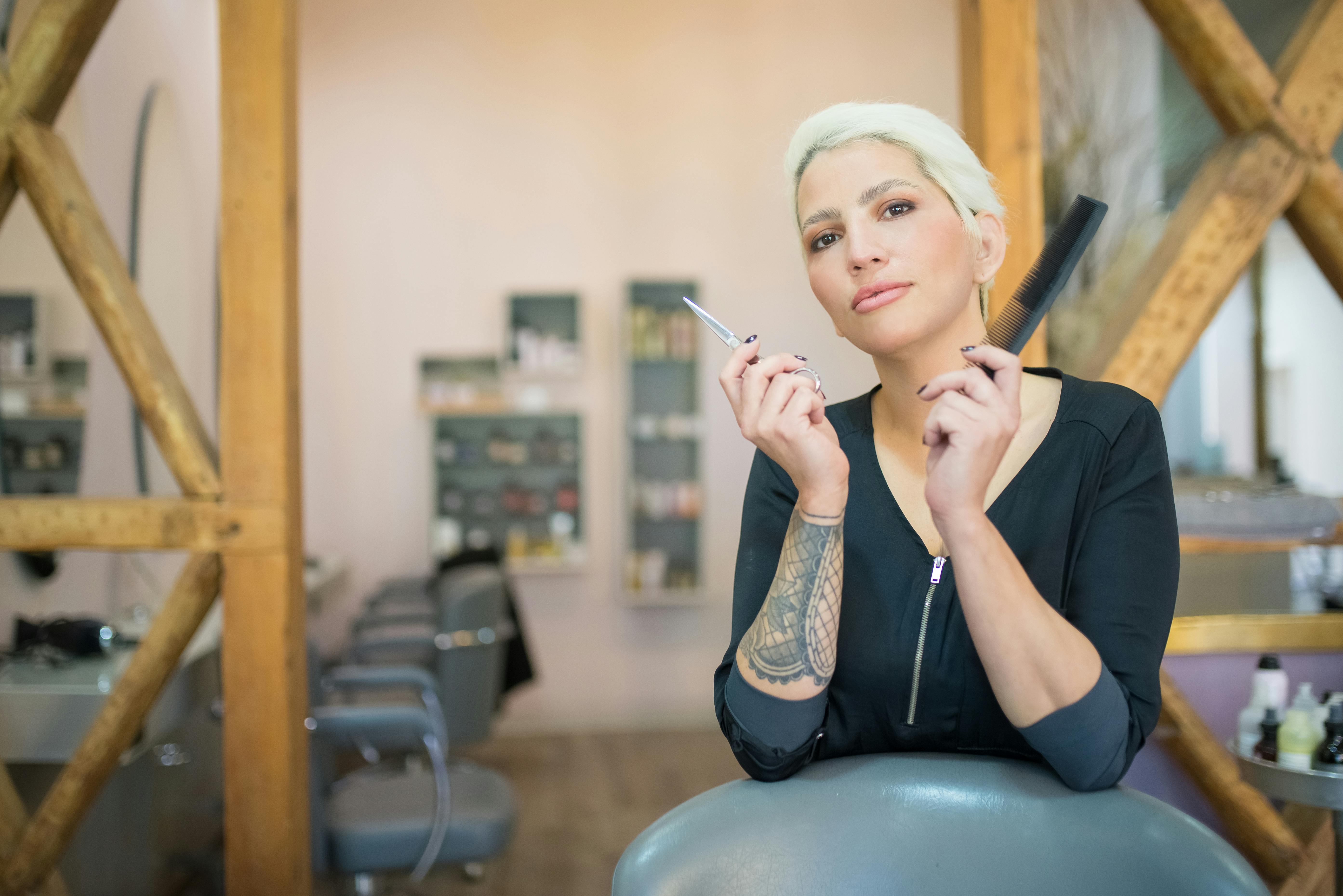 A confident hairdresser with arm tattoo and colored hair holding scissors and comb in a modern salon.