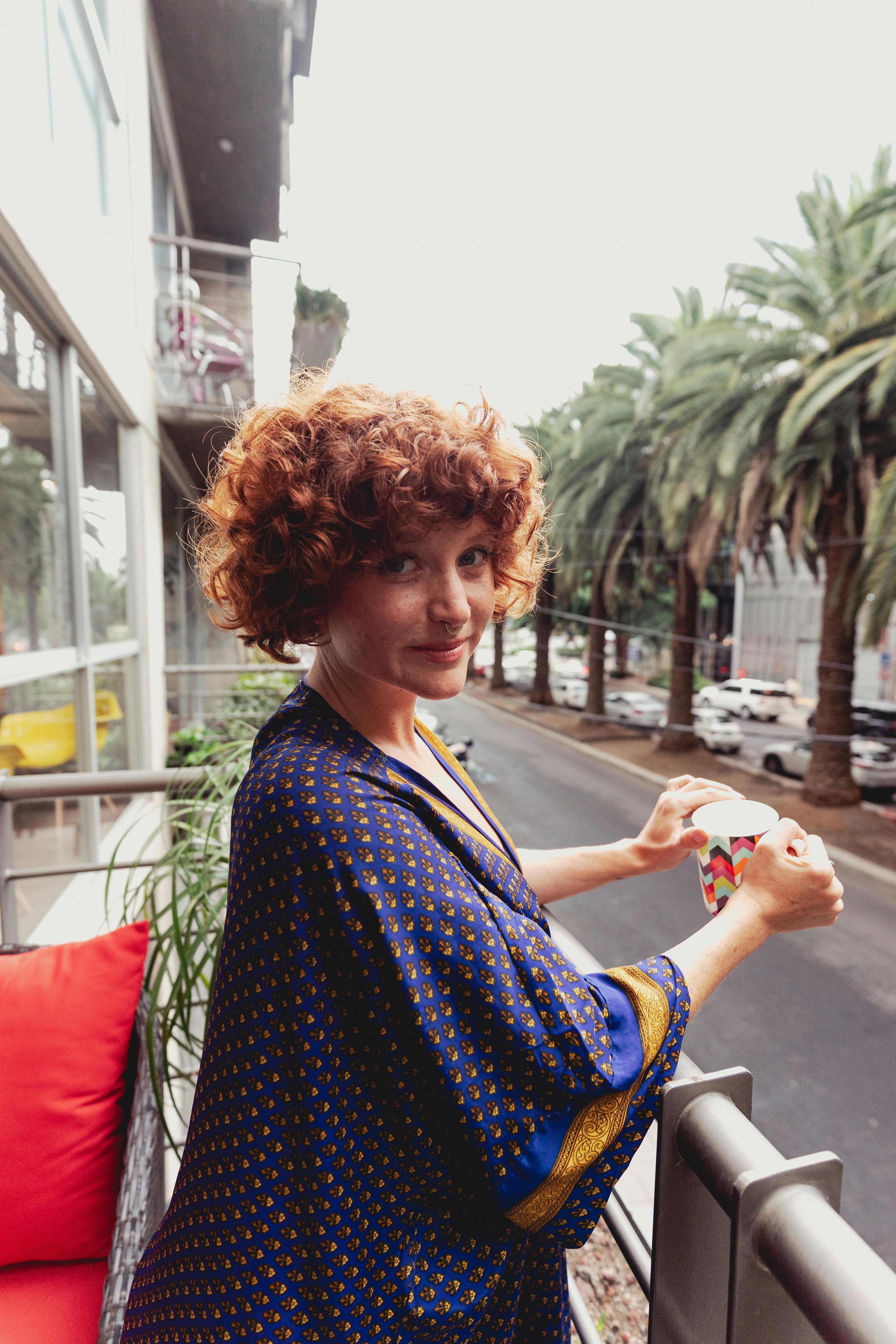 一位留着红色卷发的女人在阳台上拿着咖啡杯，俯瞰着棕榈树