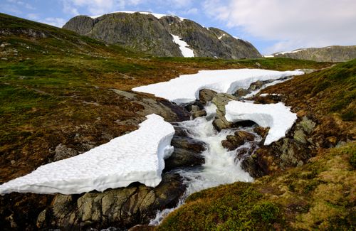 Free Capture of a serene mountain stream flowing through rocky terrain with patches of melting snow under a clear sky. Stock Photo
