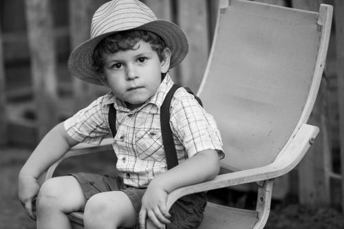 Free Cute young boy in a hat sitting on a chair, looking at the camera. Monochrome image with a vintage feel. Stock Photo