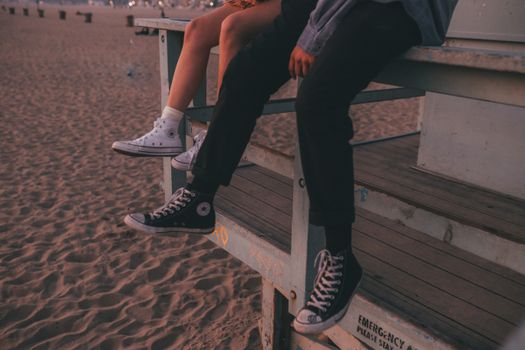 A couple sitting on a lifeguard stand at the beach wearing Converse sneakers during sunset.