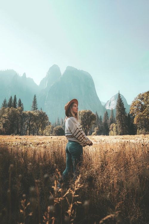 Free A woman stands in a serene countryside field with a backdrop of majestic mountains under a clear sky. Stock Photo