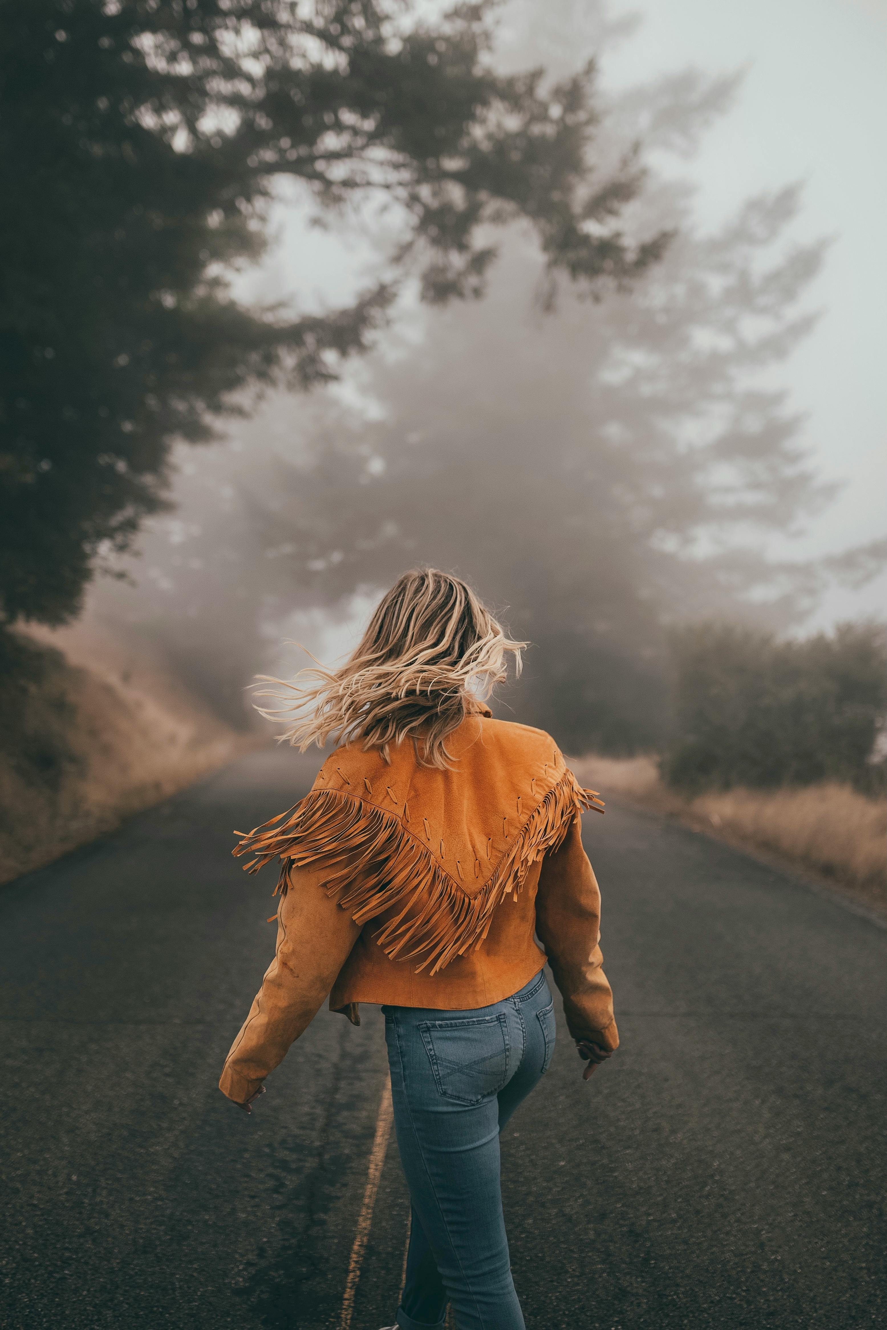 Back view of a woman walking on a foggy road surrounded by trees, wearing a brown jacket.