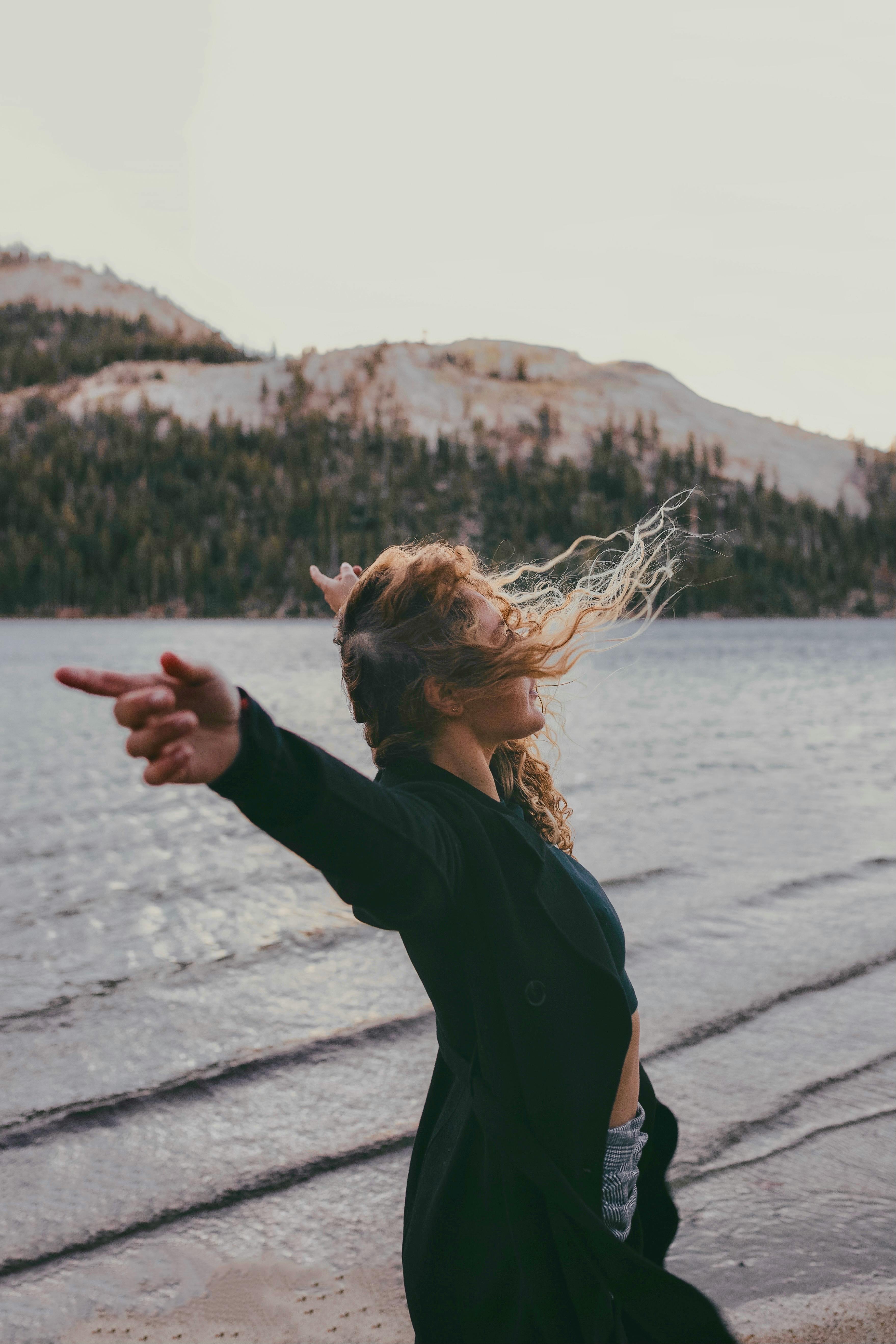 A carefree woman with blonde hair poses by a scenic mountain lake, embracing the breeze.