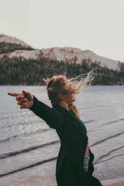 A carefree woman with blonde hair poses by a scenic mountain lake, embracing the breeze.