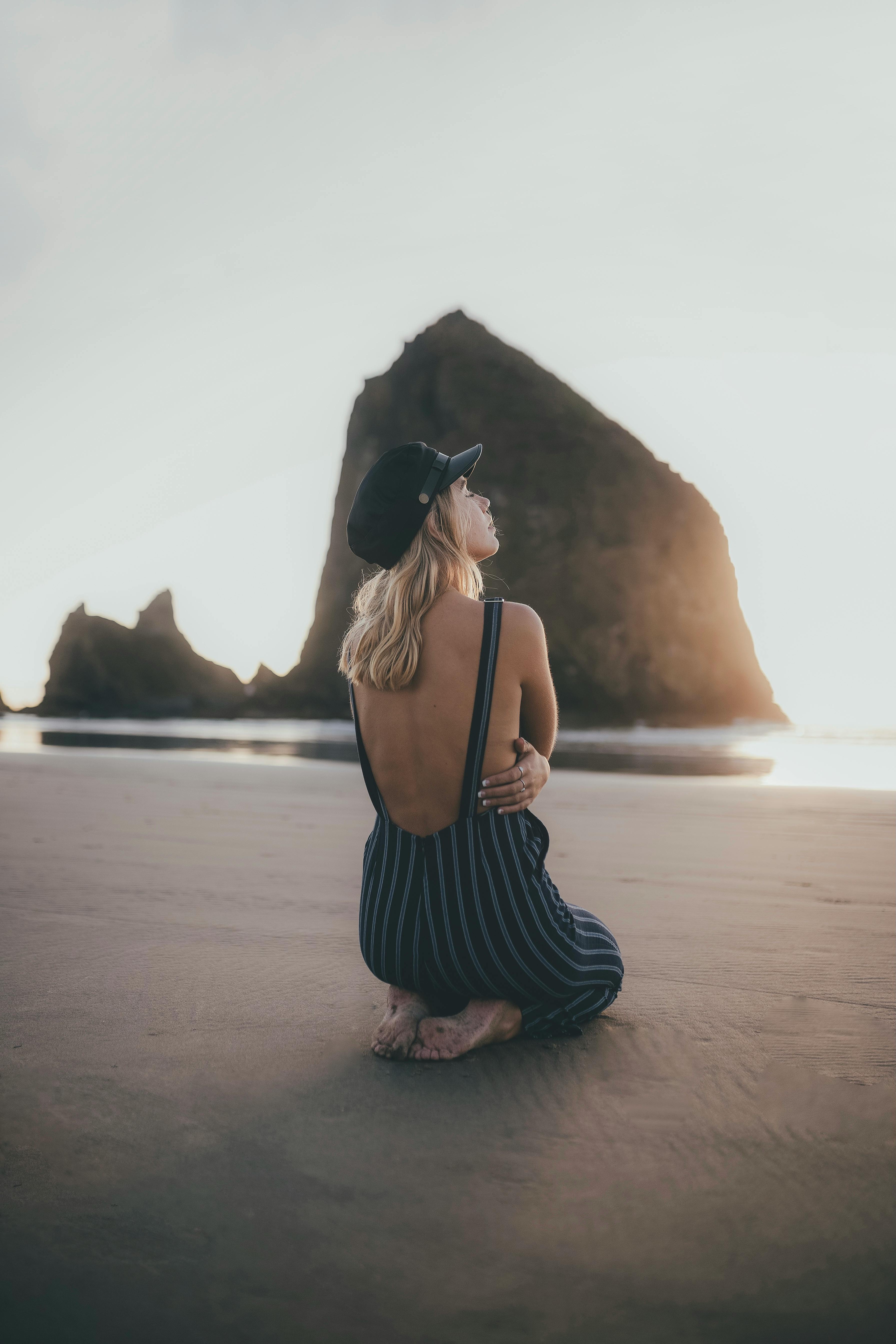 A woman kneels on a serene beach, gazing at a rock formation during sunset.