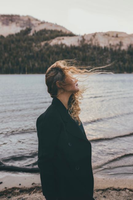 A woman in a black coat facing a windy lakeside, capturing a serene natural landscape.
