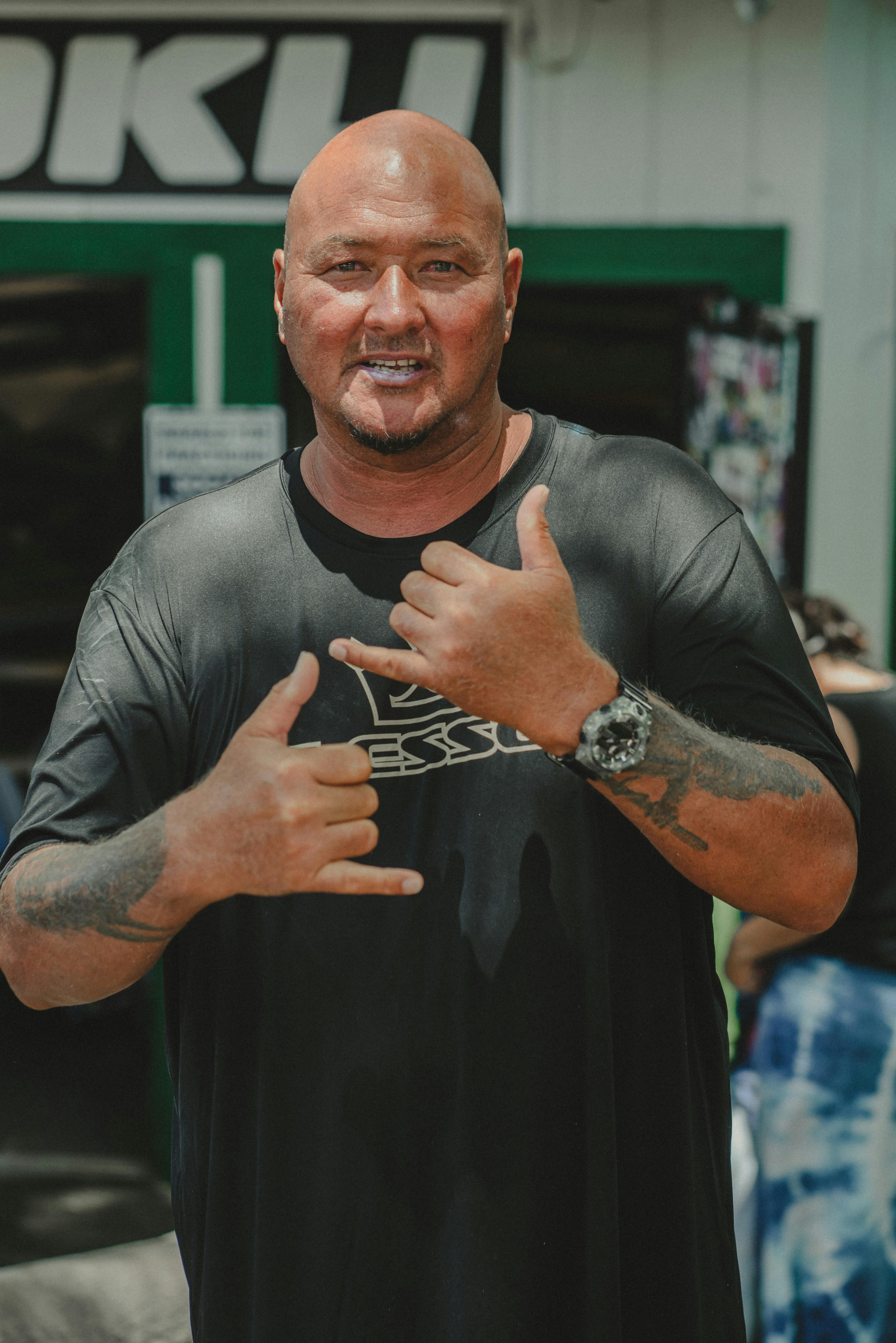 Smiling bald man making hang loose gesture in casual black shirt outdoors.