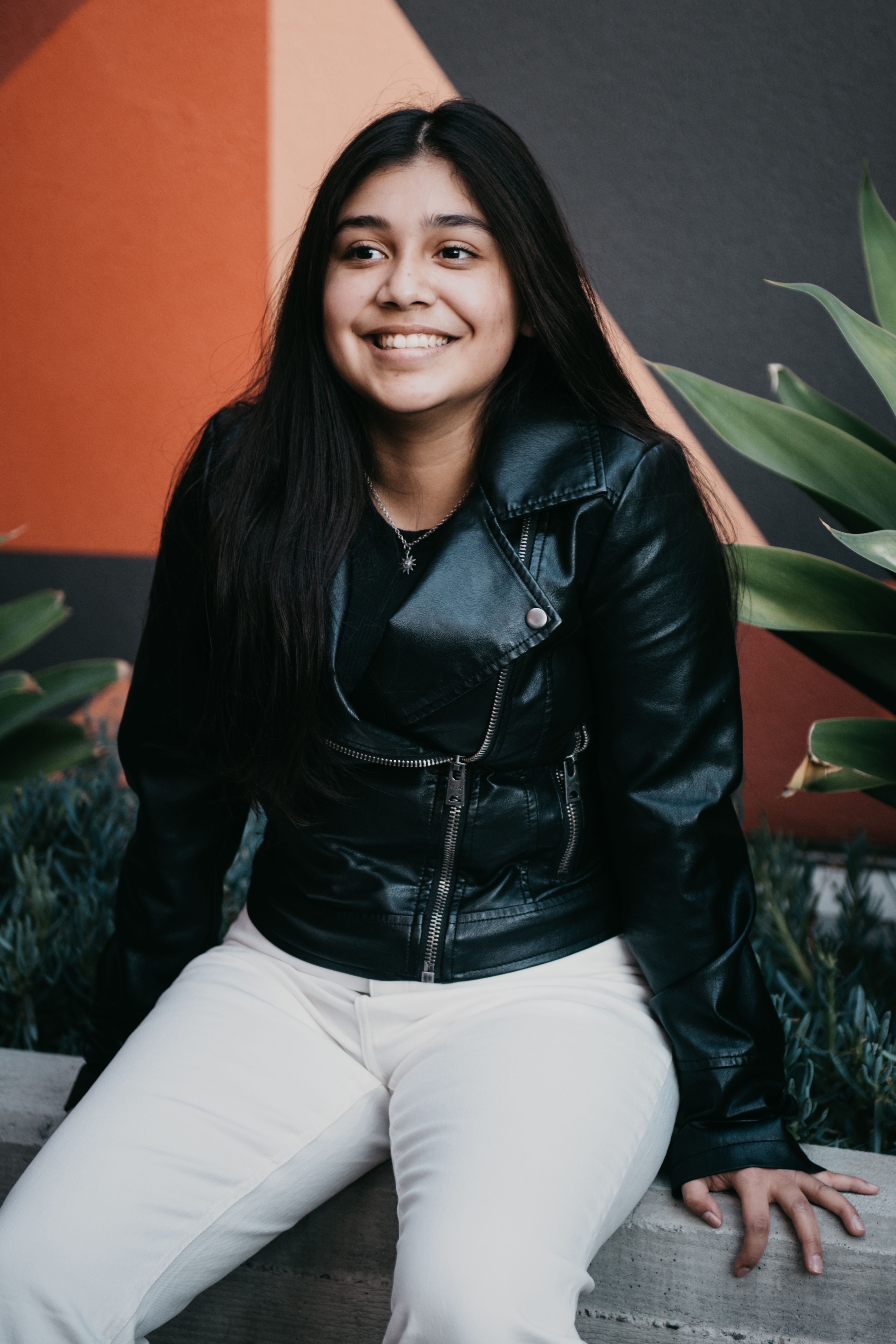 A woman in a leather jacket sits and smiles against a vibrant geometric background.
